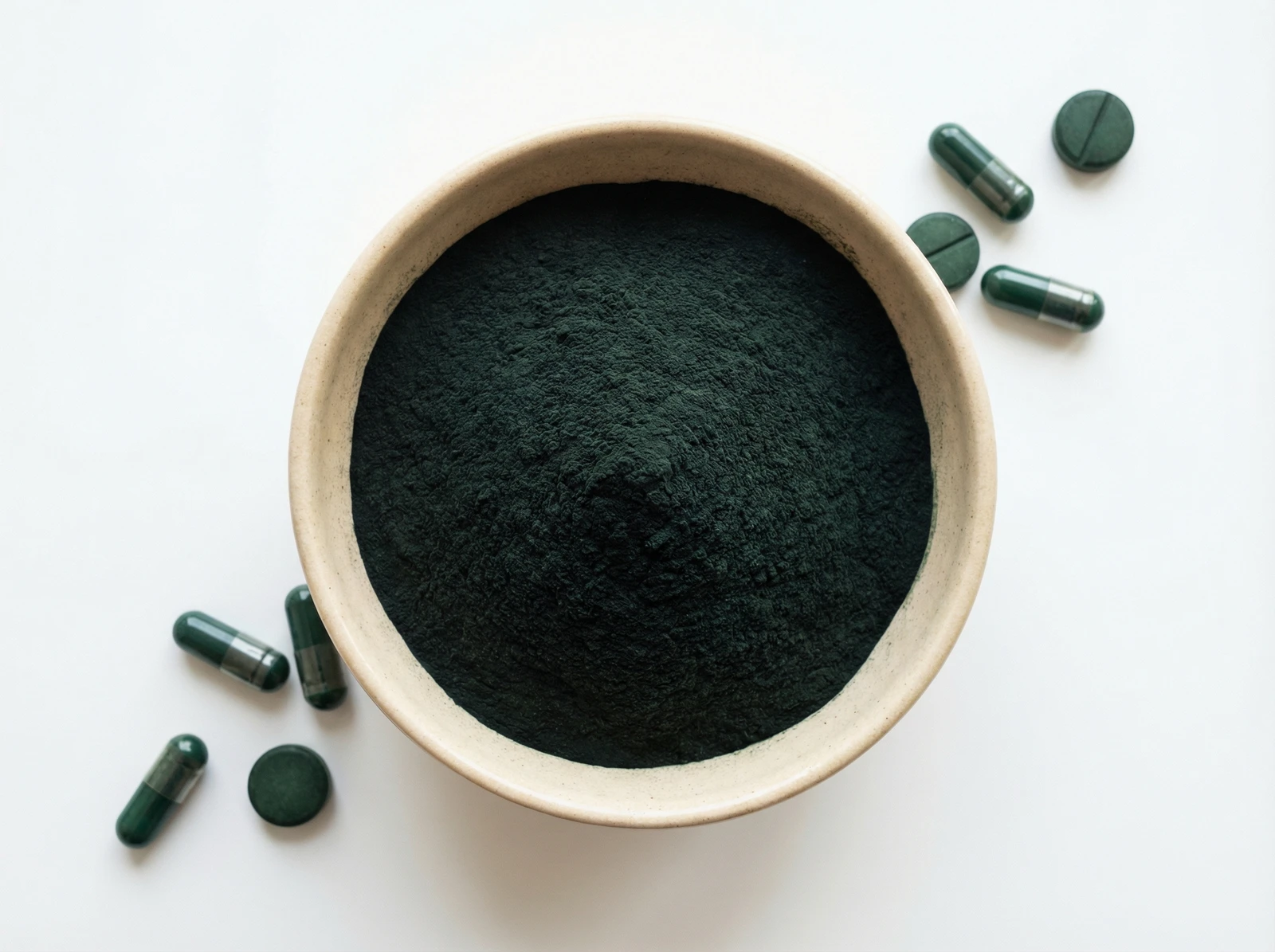 Dunkelgrünes Spirulina Pulver in einer Keramikschale, umgeben von Spirulina Kapseln und Tabletten auf weißem Hintergrund