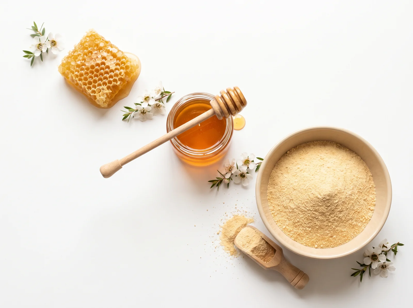 Draufsicht auf weißem Grund: links oben ein Stück goldene Honigwabe, in der Mitte ein Glas mit flüssigem Honig und einem Honiglöffel aus Holz, rechts unten eine beige Keramikschale mit hellgoldenem Honigpulver und daneben eine Holzschaufel mit Pulver. Dazwischen liegen kleine weiße Manukablüten mit grünen Blättern.